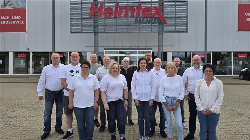 Gruppe von Menschen in weißer und heller Kleidung steht vor einem Gebäude mit dem Schriftzug 'Heimtex'