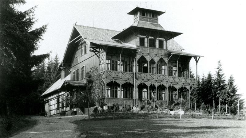 Großes historisches Fachwerkhaus mit mehreren Stockwerken und verziertem Balkon vor Nadelbäumen