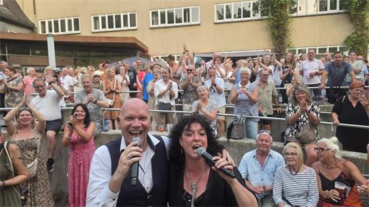 Zwei Sänger mit Mikrofonen vor einer applaudierenden Menschenmenge im Freien vor einem Gebäude