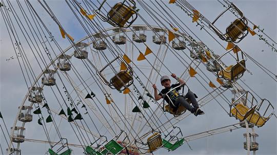 Person sitzt in einer gelben Gondel eines Kettenkarussells, das sich vor bewölktem Himmel dreht