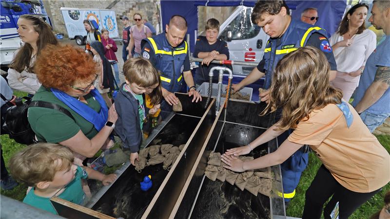 Gruppe von Kindern und Erwachsenen um einen langen, rechteckigen Wasserbehälter mit Sandsäcken unter einem blauen Pavillon.