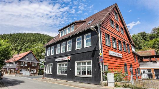 Fachwerkhaus mit schwarzer und roter Holzfassade an einer Straßenecke vor bewaldetem Hang.