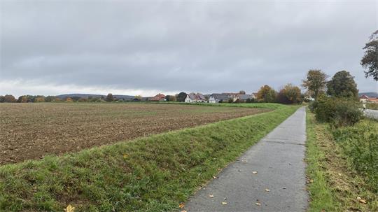 Landschaft mit gepflügtem Feld links, schmalem Asphaltweg rechts und Häusern am Horizont unter bewölktem Himmel
