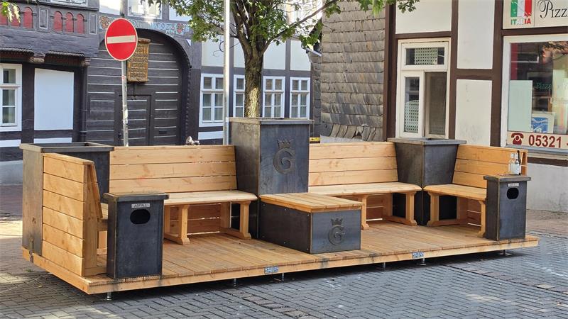 Holzplattform mit Sitzbank und Tisch aus Holz und Metall in einer Straßenszene mit Fachwerkhäusern und Verkehrsschild im Hintergrund