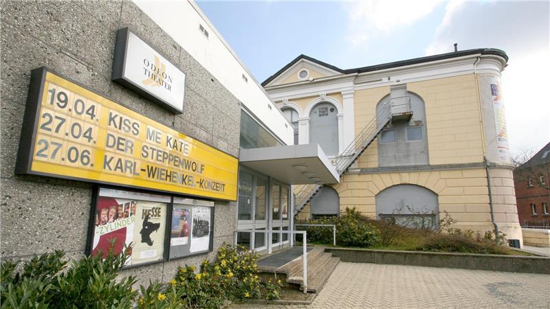 Außenansicht eines Theaters mit gelber Anzeigetafel, auf der Veranstaltungstermine stehen, und einem historischen Gebäude im Hintergrund