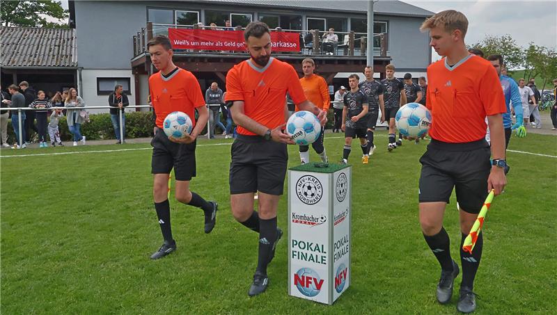 Drei Schiedsrichter in orangefarbenen Trikots tragen Fußbälle auf einem Fußballfeld, im Hintergrund Zuschauer und Spieler in schwarzen Trikots vor einem Gebäude mit Balkon