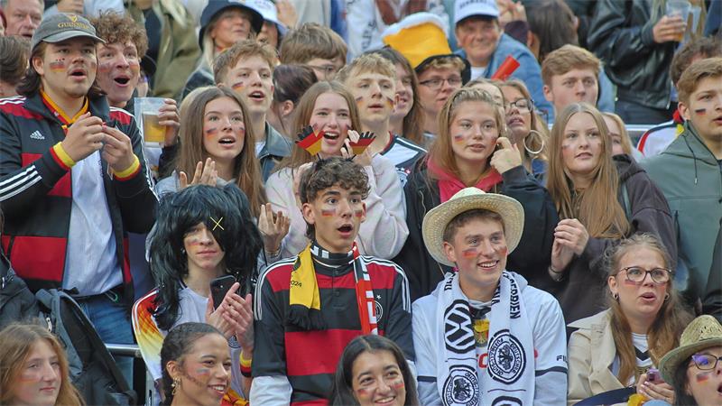 Gruppe von Menschen in einem Stadion, viele tragen schwarz-rot-gelbe Kleidung und Accessoires, einige klatschen und halten Getränke.