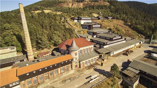 Luftaufnahme eines historischen Industriekomplexes mit Schornstein, mehreren Gebäuden und bewaldeten Hügeln im Hintergrund