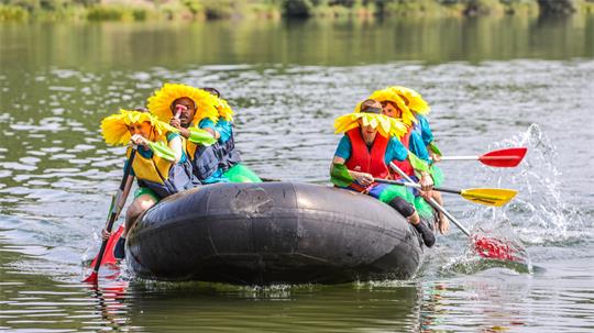 Menschen in bunten Kostümen und mit Sonnenblumenhüten paddeln mit einem Schlauchboot über einen See.