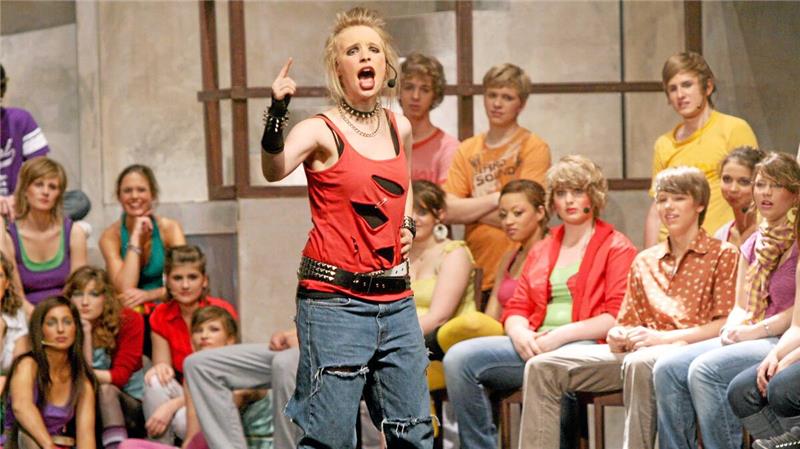 Person mit rotem ärmellosem Oberteil und zerrissener Jeanshose steht vor einer Gruppe sitzender Menschen in einem Innenraum