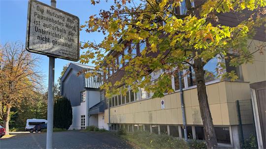 Straßenszene mit herbstlich belaubtem Baum, gelblichem Gebäude und Verkehrsschild mit schwer lesbarem Text.