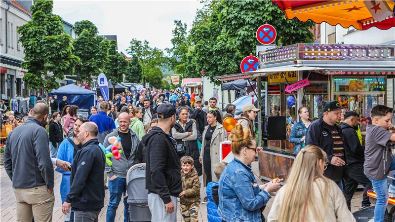 Menschenmenge auf einer belebten Straße mit Marktständen und Bäumen im Hintergrund
