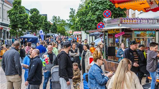 Menschenmenge auf einer belebten Straße mit Marktständen und Bäumen im Hintergrund