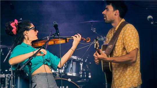 Zwei Musiker in flippigem Outfit spielen auf einer Bühne Geige und Gitarre.