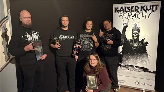 Fünf Personen in dunkler Kleidung posieren mit Pommesgabel vor einem Banner mit der Aufschrift 'KAISER KULT & KRACH!' und einer Skelettfigur mit erhobener Hand.