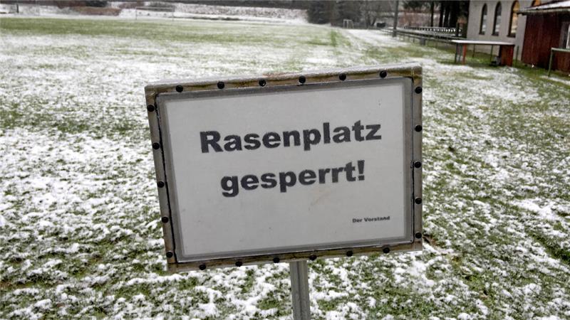 Schild mit der Aufschrift 'Rasenplatz gesperrt' auf einem schneebedeckten Sportplatz mit Wald im Hintergrund