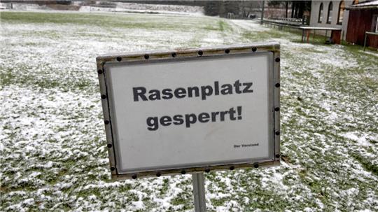 Schild mit der Aufschrift 'Rasenplatz gesperrt' auf einem schneebedeckten Sportplatz mit Wald im Hintergrund