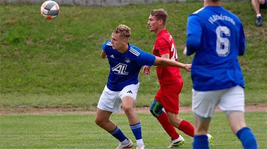 Zwei Fußballspieler in Aktion auf dem Spielfeld, einer in blauem Trikot und weißer Hose, der andere in rotem Trikot, im Hintergrund ein weiterer Spieler in blauem Trikot mit der Nummer 6.