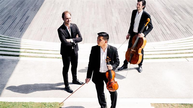 Drei Männer in Anzügen auf einer hellen Treppe, einer hält eine Violine, einer ein Cello, der dritte steht mit verschränkten Armen