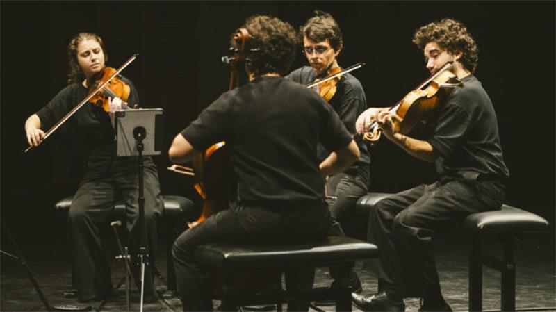Vier Musiker sitzen im Halbkreis auf Stühlen und spielen Geige auf einer dunklen Bühne.