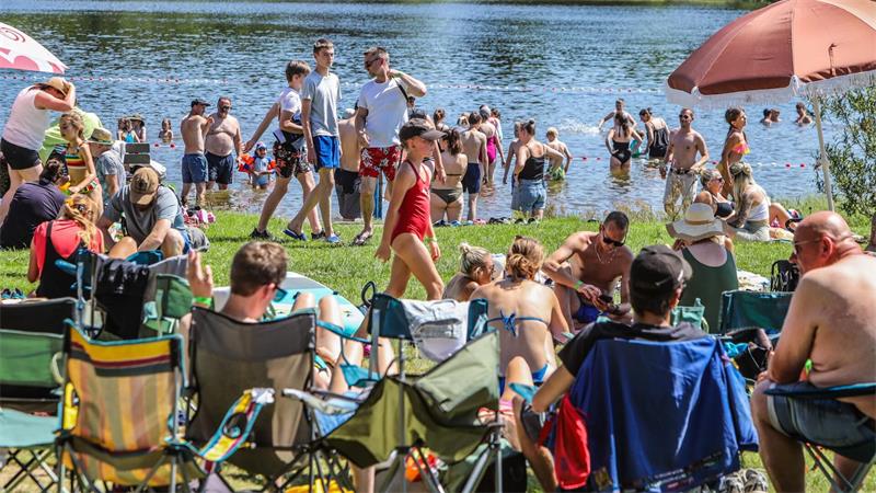 Menschen sitzen auf Campingstühlen und liegen auf Decken am Ufer, während weitere Personen im Wasser baden.