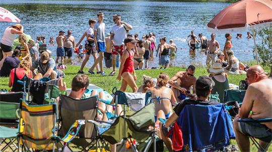 Menschen sitzen auf Campingstühlen und liegen auf Decken am Ufer, während weitere Personen im Wasser baden.