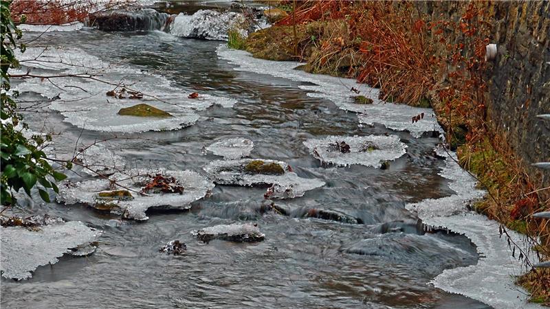 Fluss mit teilweise zugefrorenen Stellen und kahlen Büschen am Ufer neben einer Steinmauer.