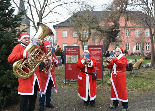 Vier Musiker in Weihnachtsmannkostüm spielen in einem Außengelände auf Blasinstrumenten.