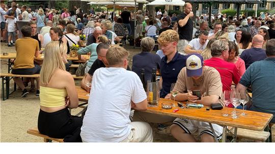 Ein Festgelände. Zahlreiche Menschen sitzen an Tischen, unterhalten sich und trinken.