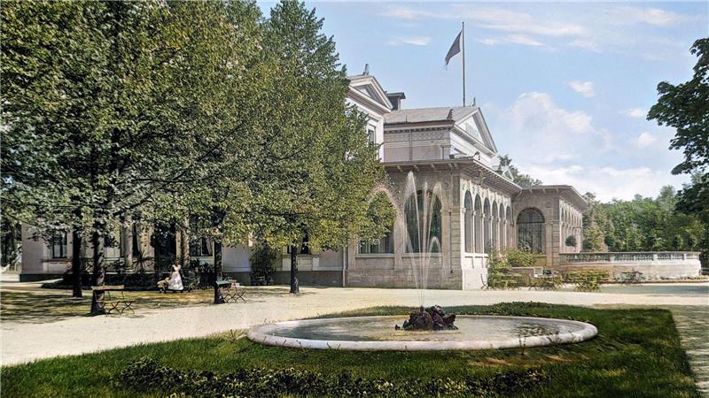 Zusehen ist ein Brunnen in einem Garten und im Hintergrund ist ein weißes Gebäude mit einer Flagge auf dem Dach.