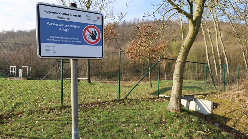 Schild mit Verbotssymbol und Text vor eingezäuntem, grasbewachsenem Gelände mit Bäumen und einem kleinen Betonkasten