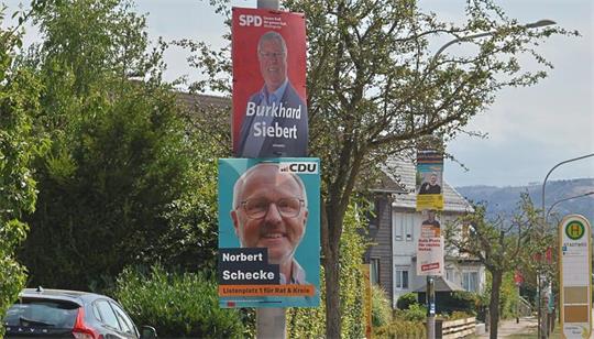 Straße mit mehreren Wahlplakaten an Laternenmasten, im Vordergrund Plakate von Burkhard Siebert (SPD) und Norbert Schecke (CDU)