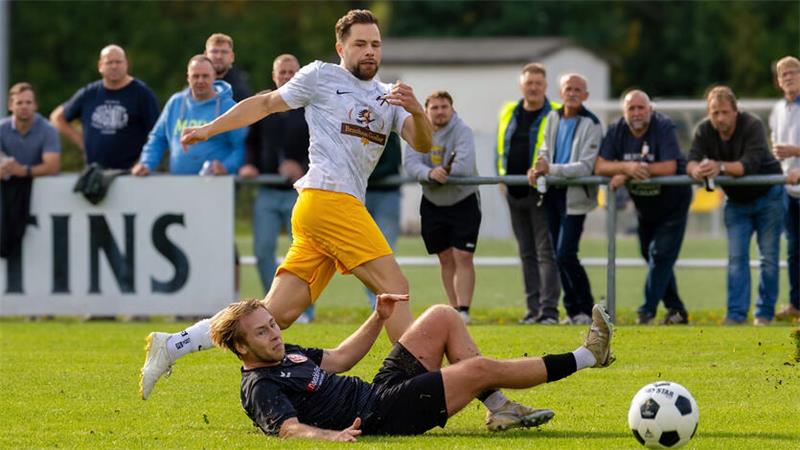 Fußballspieler in gelber Hose und weißem Trikot schießt den Ball, am Boden liegende ein Spieler in schwarzer Kleidung auf einem Spielfeld mit Zuschauern im Hintergrund