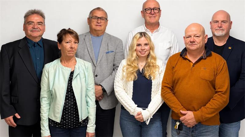 Gruppe von sieben Personen unterschiedlichen Alters steht vor weißem Hintergrund, drei Frauen und vier Männer, alle blicken frontal in die Kamera