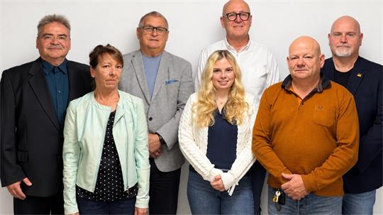 Gruppe von sieben Personen unterschiedlichen Alters steht vor weißem Hintergrund, drei Frauen und vier Männer, alle blicken frontal in die Kamera