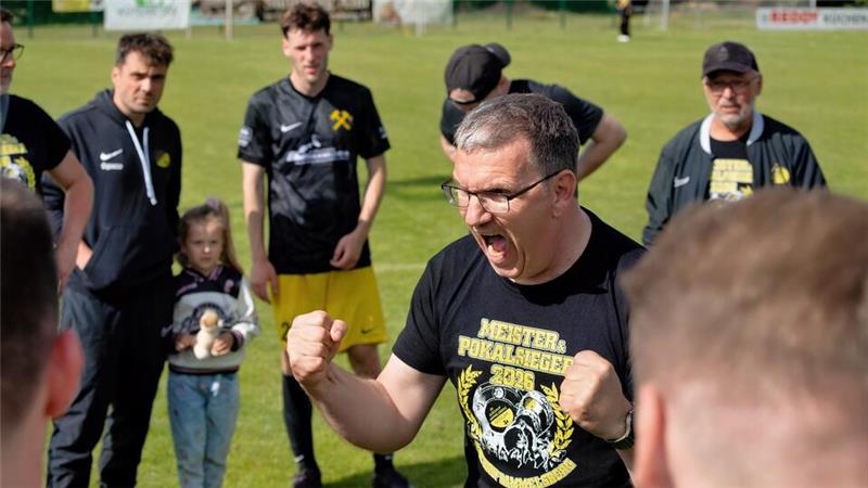 Mann mit erhobenen Fäusten und schwarzem T-Shirt mit Aufdruck 'MEISTER POKALSIEGER 2016' umgeben von mehreren Personen auf einem Fußballfeld.