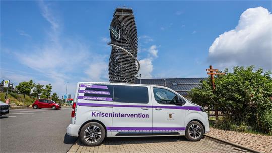 Weißer Transporter mit lila Schrift 'Krisenintervention' steht vor einem modernen, spiralförmigen Turm unter blauem Himmel mit Wolken.