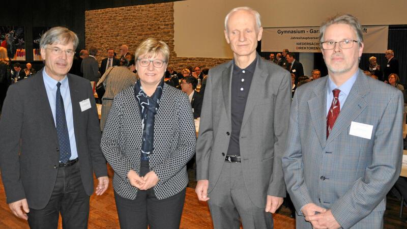 Vier Personen in formeller Kleidung stehen in einem Innenraum mit weiteren Personen im Hintergrund