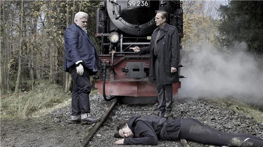 Ein weibliche Leiche liegt auf einem Bahngleis, dahinter ist eine Dampflok zu sehen. Außerem unterhalten sich zwei Männer.