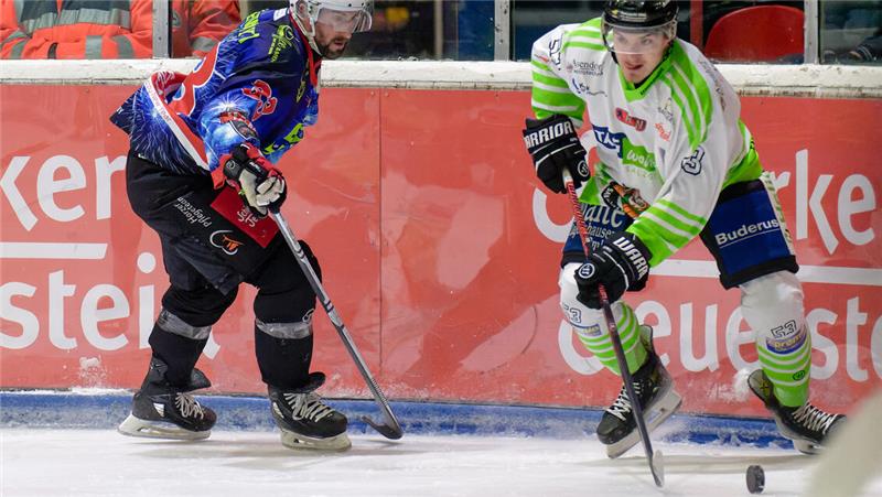 Zwei Eishockeyspieler in Aktion, einer in blau-roter Ausrüstung, der andere in weiß-grüner Ausrüstung, beide mit Schlägern und Schlittschuhen auf dem Eis.