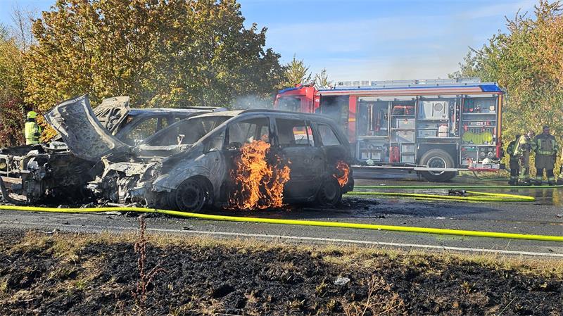 Zwei komplett ausgebrannte Fahrzeuge stehen auf der Straße. Ein Feuerwehrfahrzeug steht dahinter.