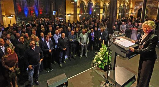Große Menschenmenge in einem prunkvollen Saal mit hohen, verzierten Bögen, eine Frau steht am Rednerpult mit Blumenarrangement davor.