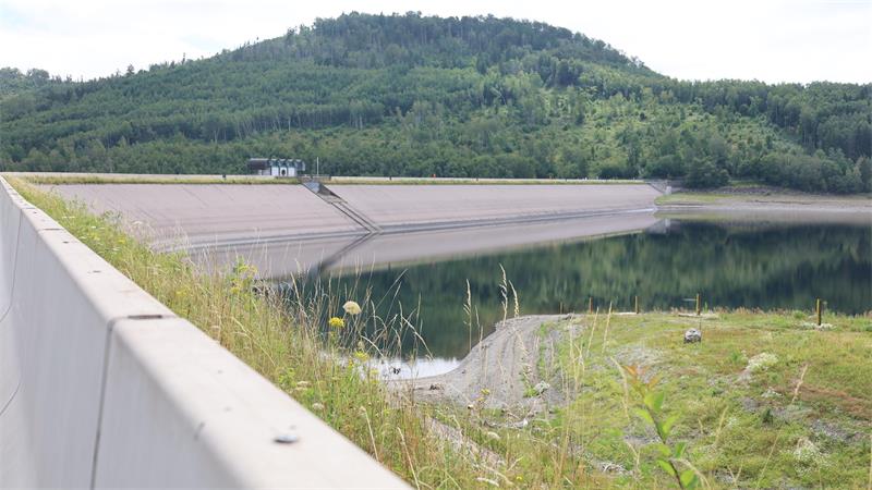 Stausee mit Betonmauer im Vordergrund und bewaldetem Hügel im Hintergrund
