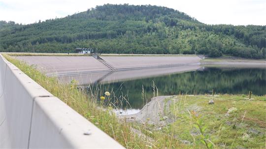 Stausee mit Betonmauer im Vordergrund und bewaldetem Hügel im Hintergrund