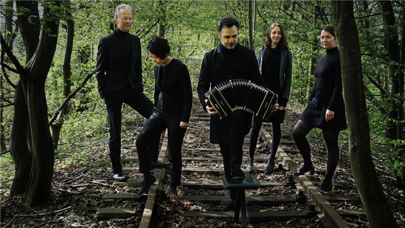 Fünf schwarz gekleidete Menschen stehen locker verteilt im Wald auf einem Bahngleis. Der Frontmann hält ein geöffnetes Akkordeon in Händen.