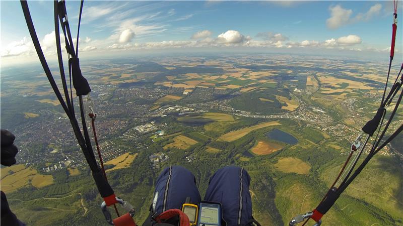 Blick aus der Vogelperspektive beim Gleitschirmfliegen auf eine Landschaft mit Feldern, Wäldern, Seen und Ortschaften unter blauem Himmel mit Wolken.