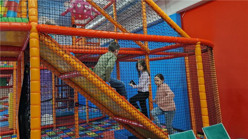 Drei Kinder spielen in einem bunten Indoor-Spielplatz mit Rutsche und Netzumzäunung.