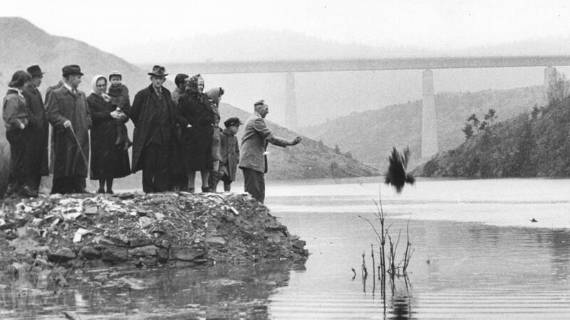Einige Bürger sehen dabei zu, wie Bürgermeister Böhm den Strauß in die Talsperre wirft.