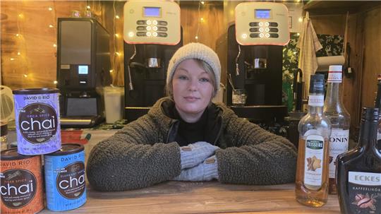 Person mit grauer Mütze und Handschuhen lehnt an einem Holztresen, im Hintergrund zwei Kaffeeautomaten und verschiedene Getränkedosen und Flaschen.