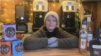 Person mit grauer Mütze und Handschuhen lehnt an einem Holztresen, im Hintergrund zwei Kaffeeautomaten und verschiedene Getränkedosen und Flaschen.